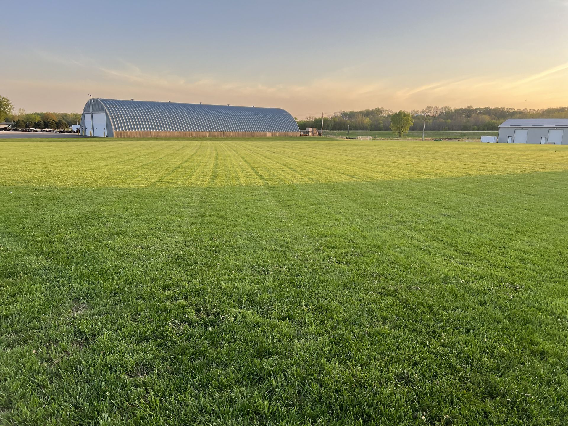 Green field with a large silver building in the background under a blue sky at sunset.