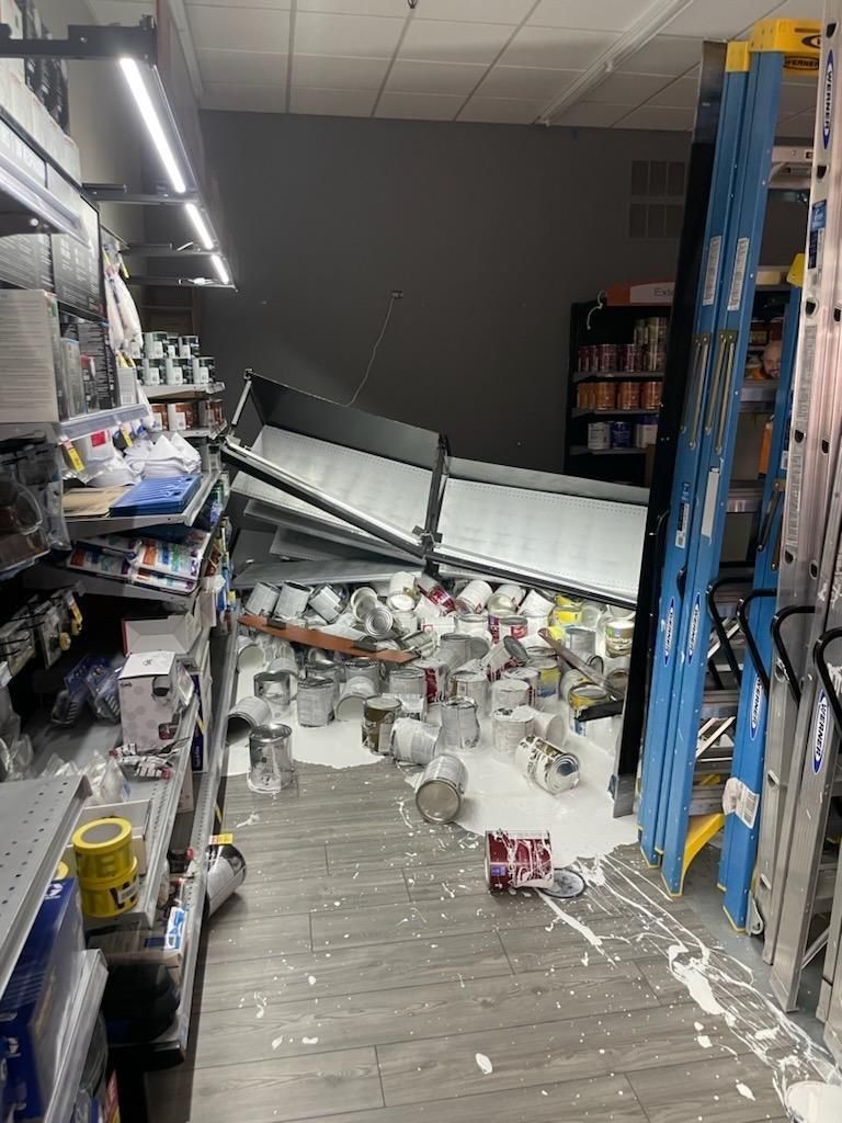 Paint cans spilled in hardware store aisle after shelving collapse. White paint covers floor.