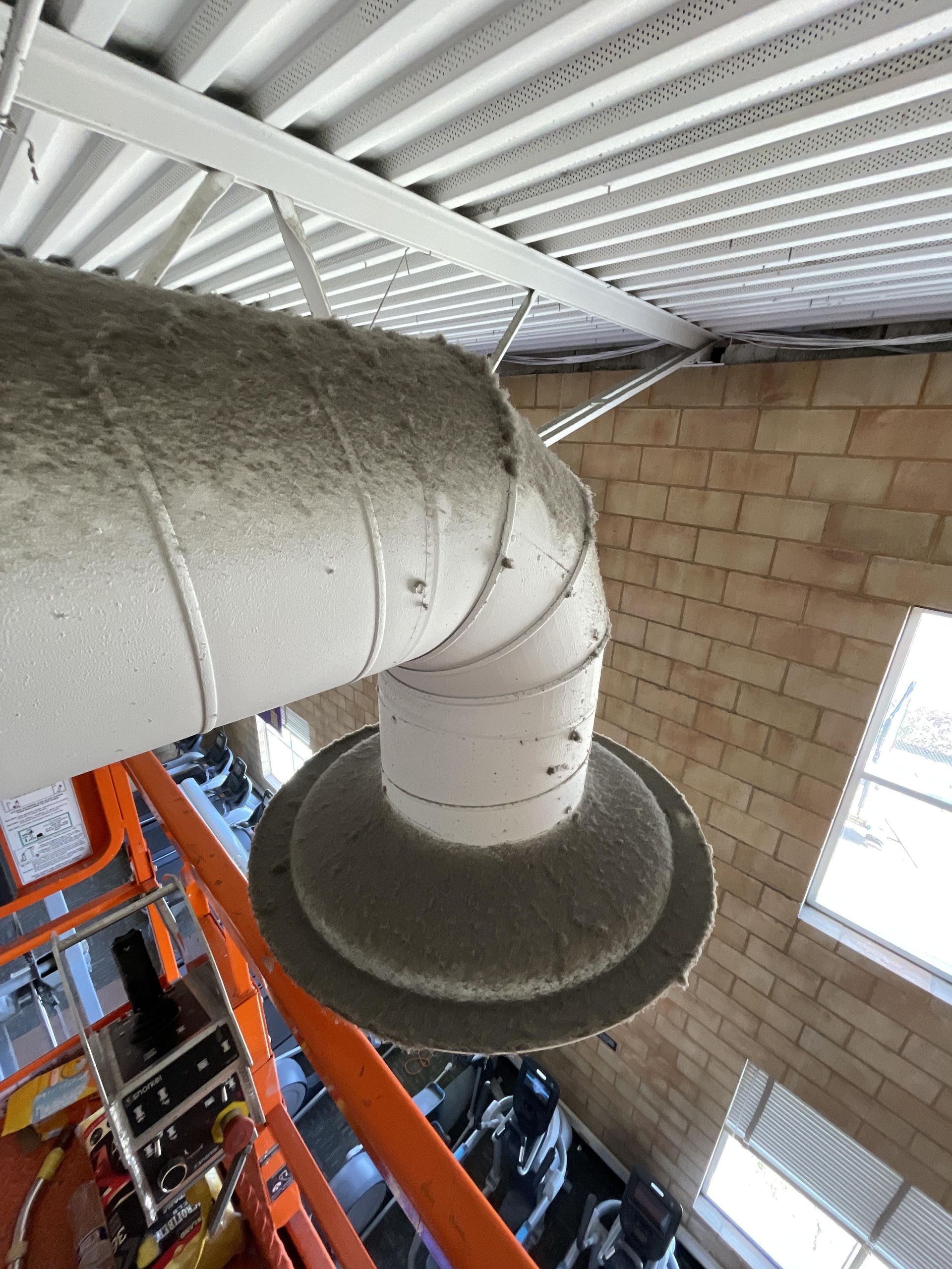Overhead view of a large, dusty ventilation duct, bent at an angle, near a brick wall and a metal ceiling.