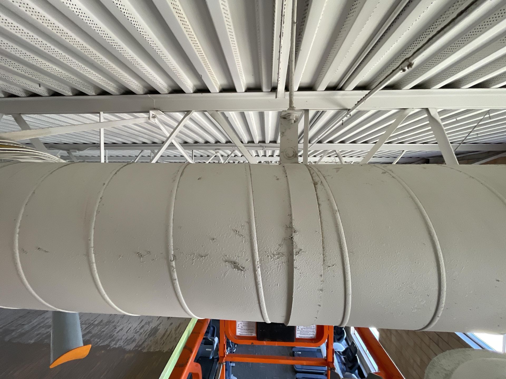 White-painted pipe with metal bands, supported by beams, under a ribbed ceiling. Orange lift below.