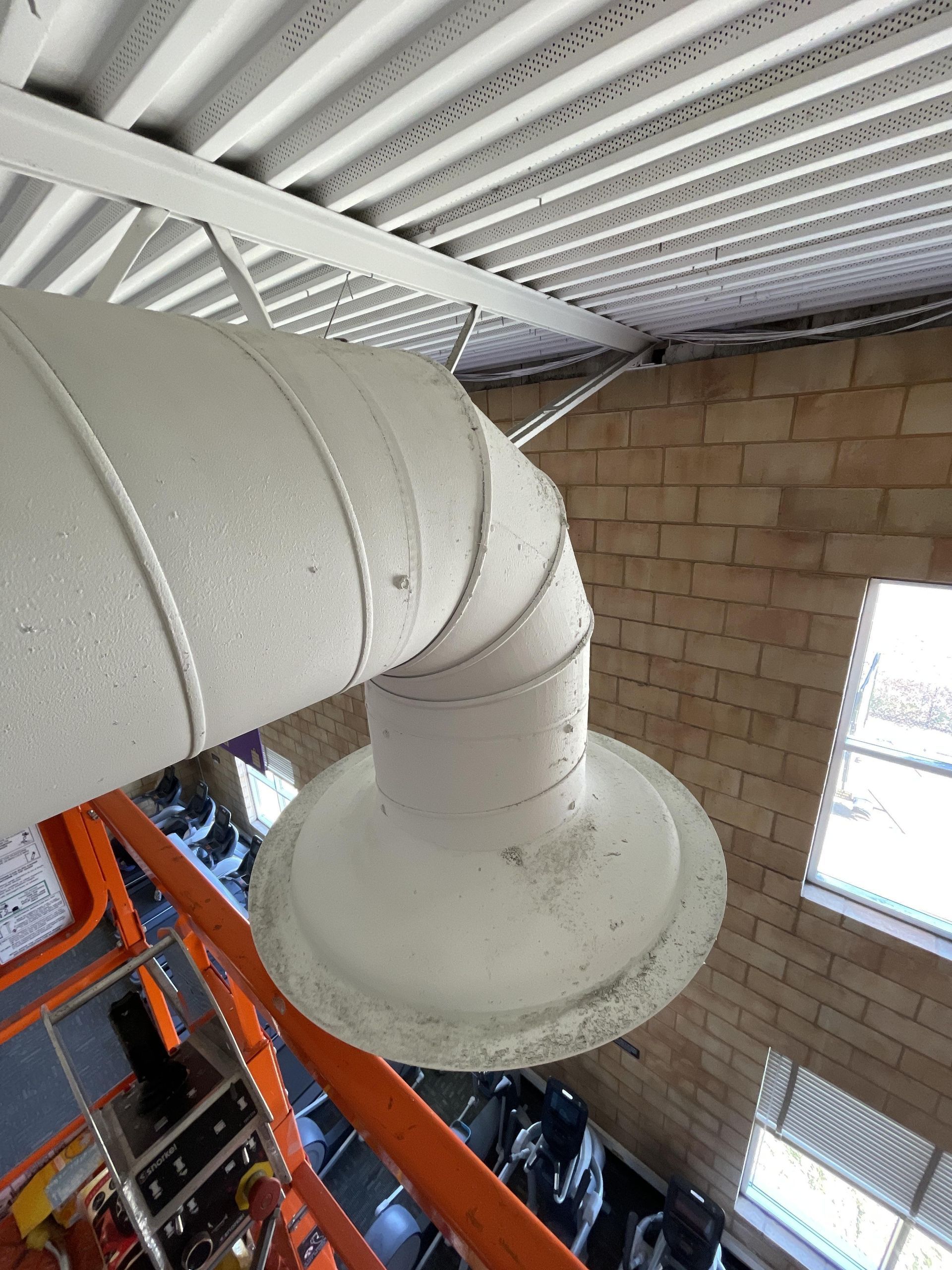 White HVAC ducting bends from ceiling near a brick wall and a window, seen from a lift.