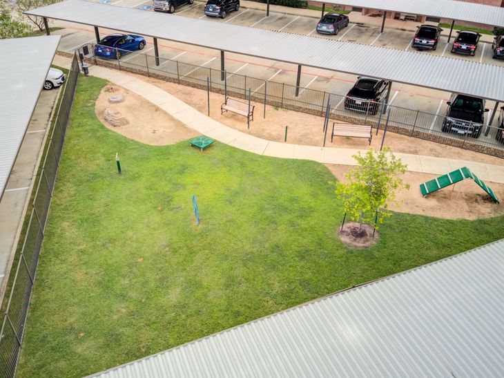 Dog park with grass, walking path, benches, and obstacles, next to a parking area.