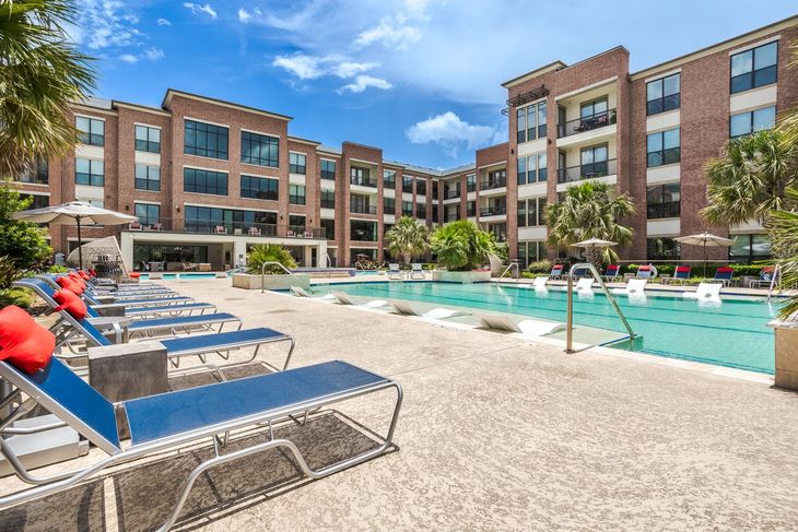 Swimming pool with lounge chairs, brick apartment building, and blue sky.