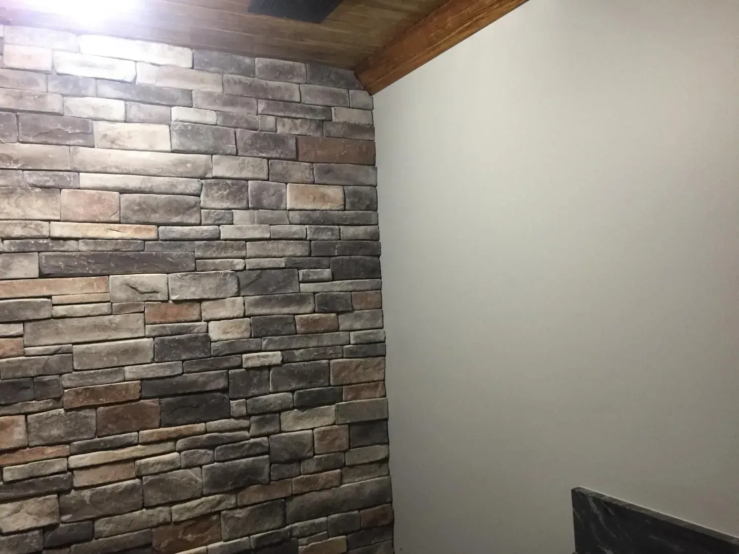 Corner of a room with a stone veneer wall and a plain, off-white painted wall. The ceiling is wood-paneled.