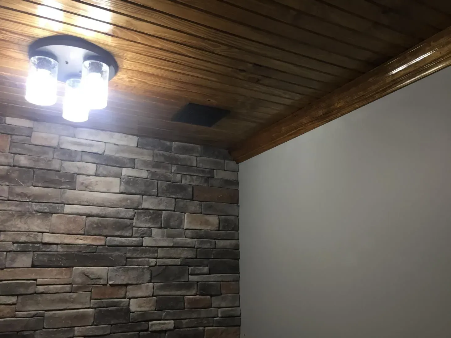 Room interior with a stone accent wall, wood ceiling, and a light fixture. The opposite wall is plain white.