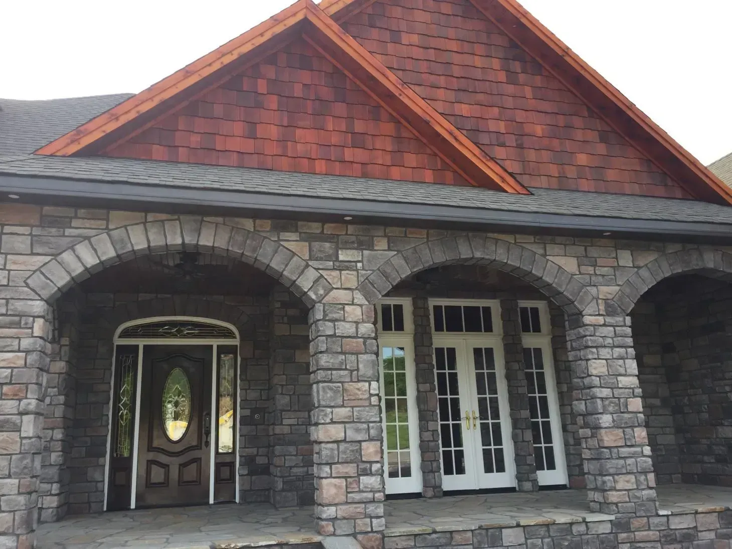 Stone facade with arched entryway and wooden front doors.  Reddish-brown shingles on the roof.