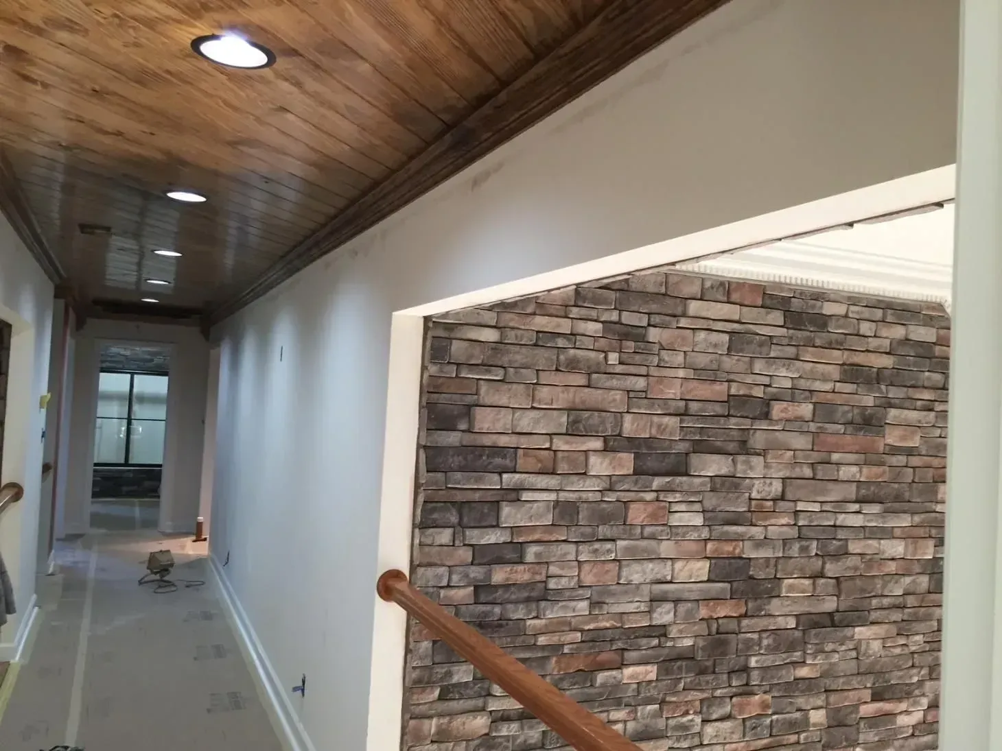 Hallway with a wooden ceiling, white walls, and a stone accent wall visible through an opening. A wooden handrail is in the foreground.