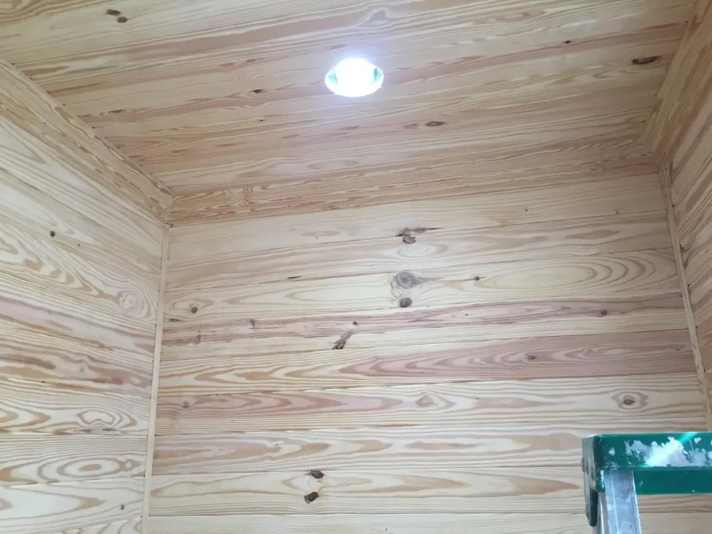 Wooden walls and ceiling of a room with a single recessed light. A green stepladder is visible in the corner.