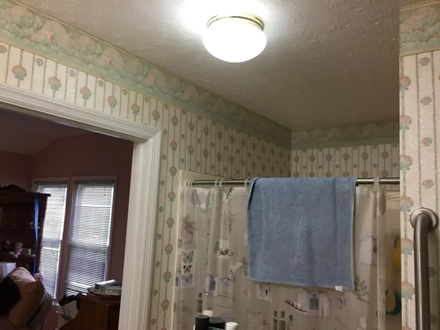 Bathroom interior with floral wallpaper, a blue towel on a shower rod, and a bright ceiling light. A doorway on the left leads to a bedroom.