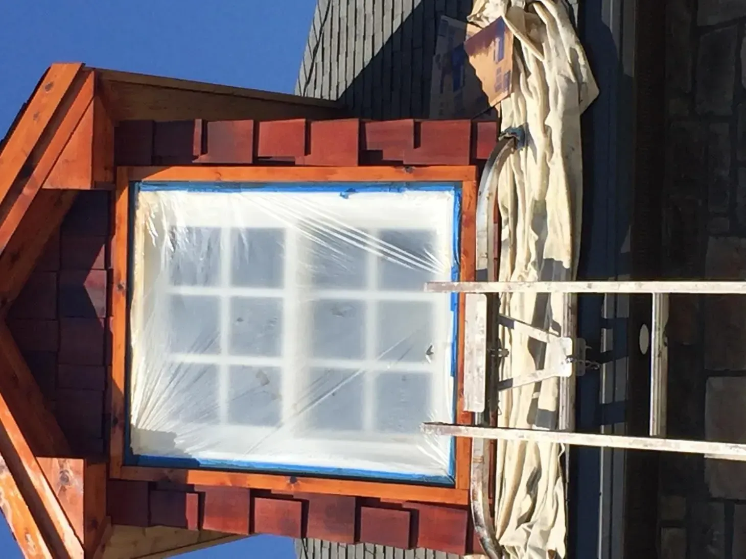 Window covered in plastic and blue tape, being painted on an exterior wooden structure. A ladder is visible.