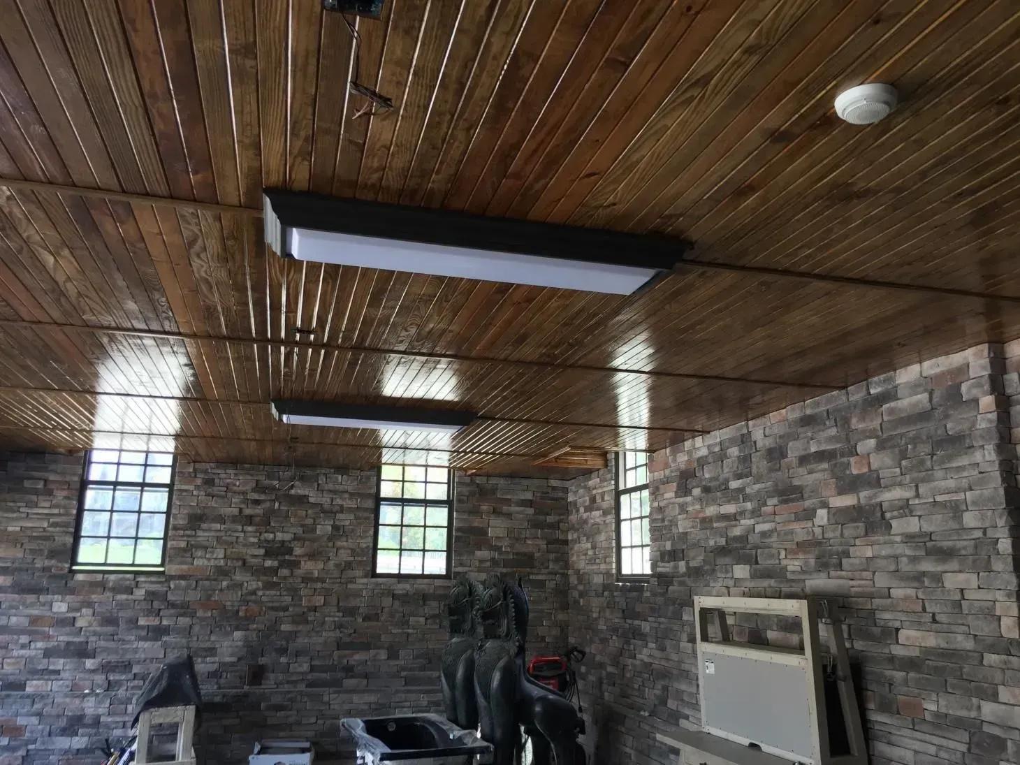 Interior view of a stone-walled room with a wooden ceiling and three rectangular lights. Windows are visible along one wall.