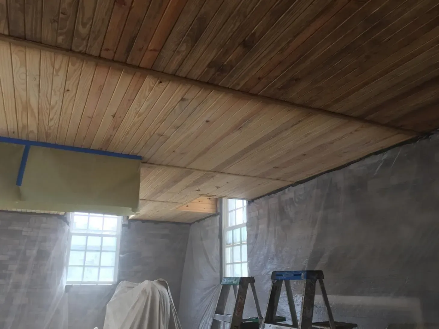 Wooden ceiling under construction in a room with gray walls, windows, and a stepladder. Blue tape and protective sheeting are visible.