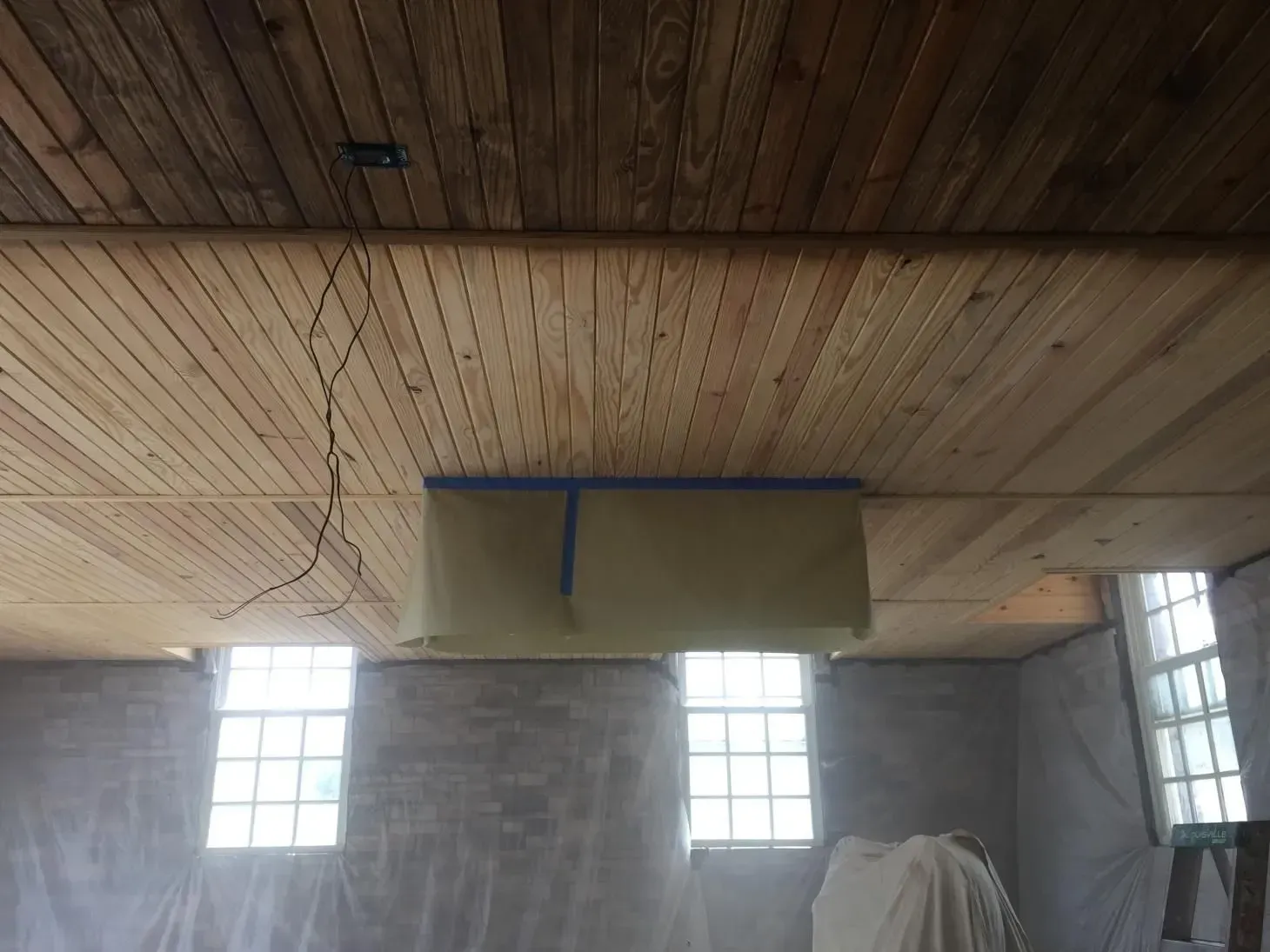 A room under renovation with a wood ceiling and three windows along one wall. A tarp is taped up to protect an area.