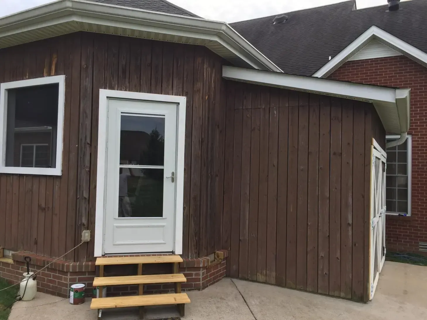 Brown wooden building with a white door and small steps, attached to a brick house. The door has a glass screen.