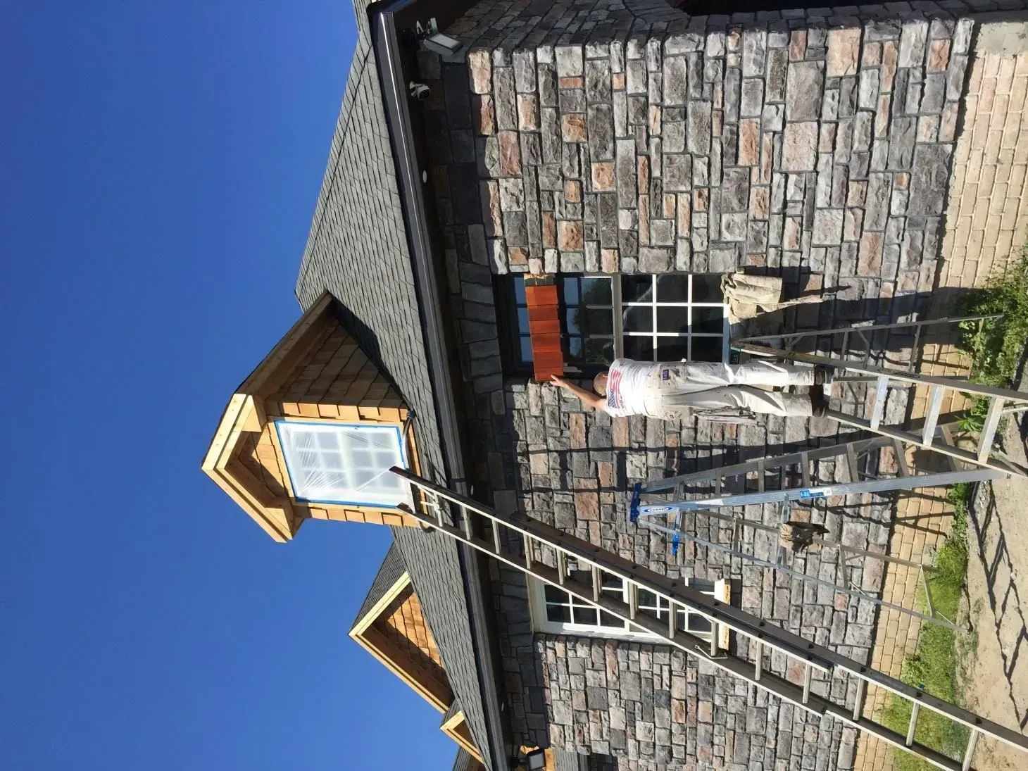 A person on a ladder painting a window of a stone-faced house. The sky is blue, and the roof is shingled.