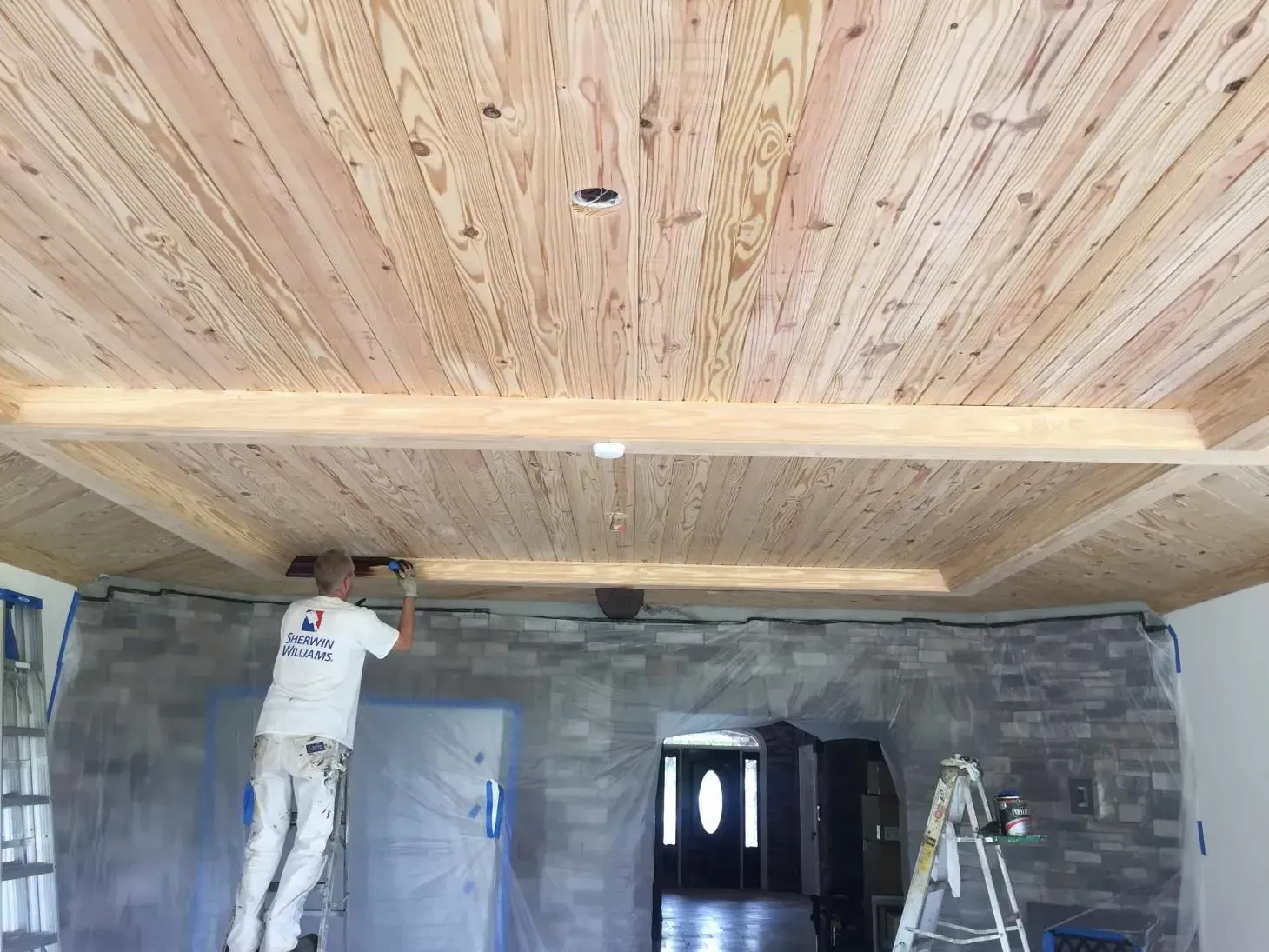 A person painting a wood ceiling with recessed lighting, a stone wall, and doorway in the background.