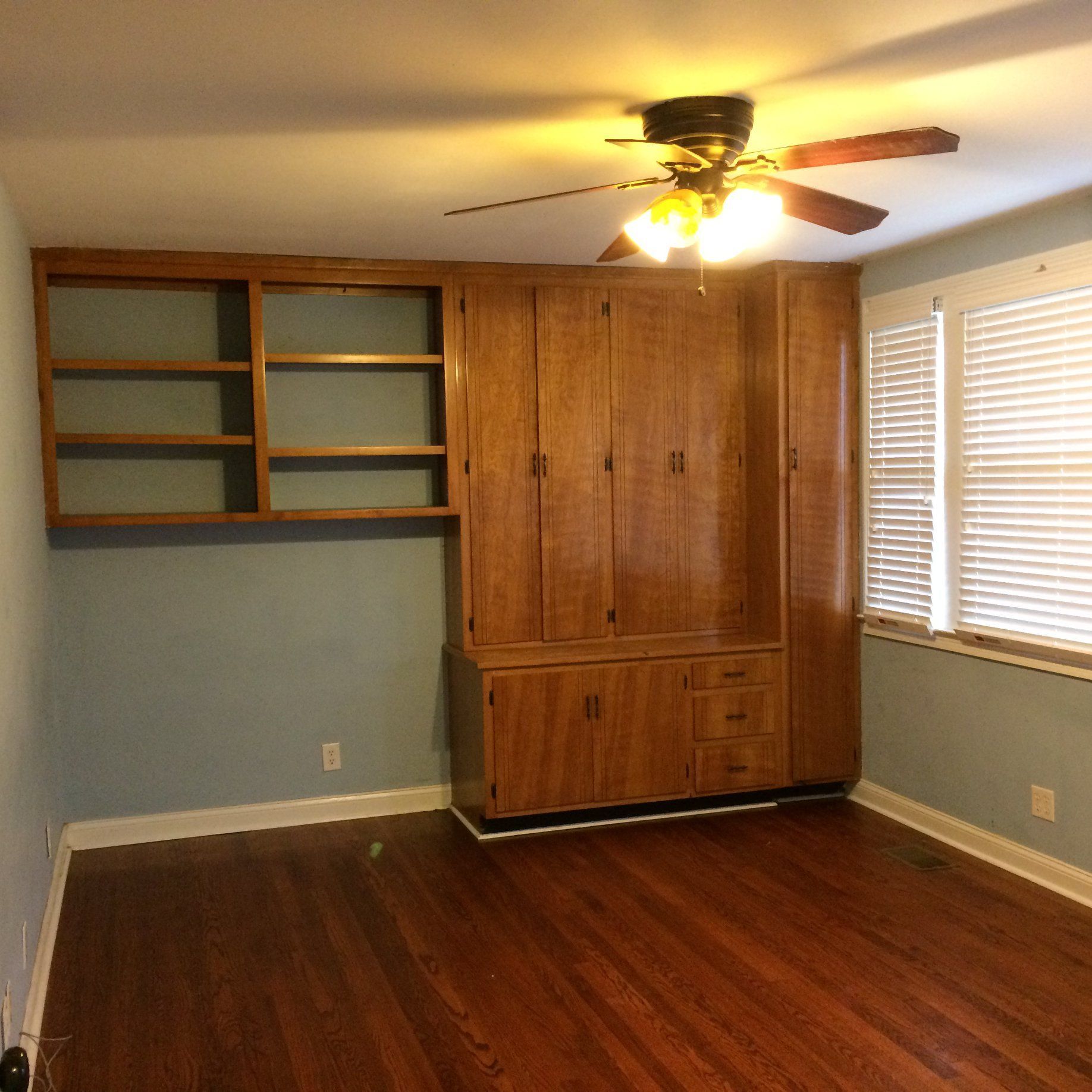 Empty bedroom with wood cabinets, built-in shelving, hardwood floors, and a window with white blinds.