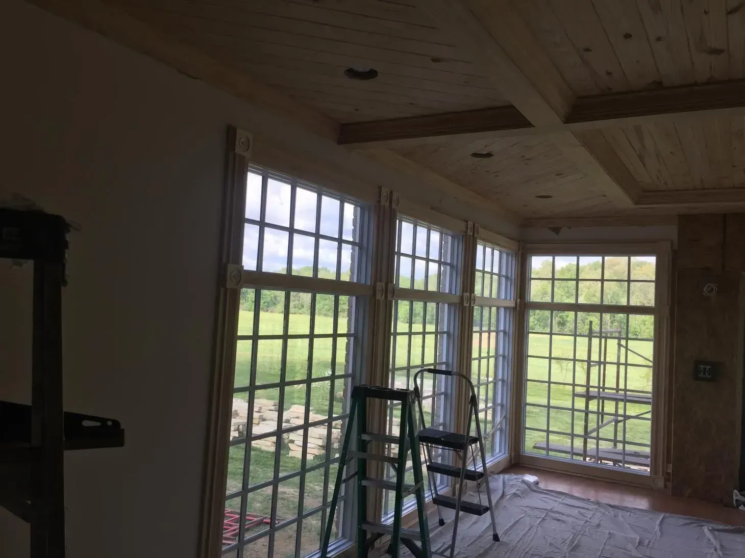 Interior room with large grid windows overlooking a green field; a stepladder stands in front of the windows.