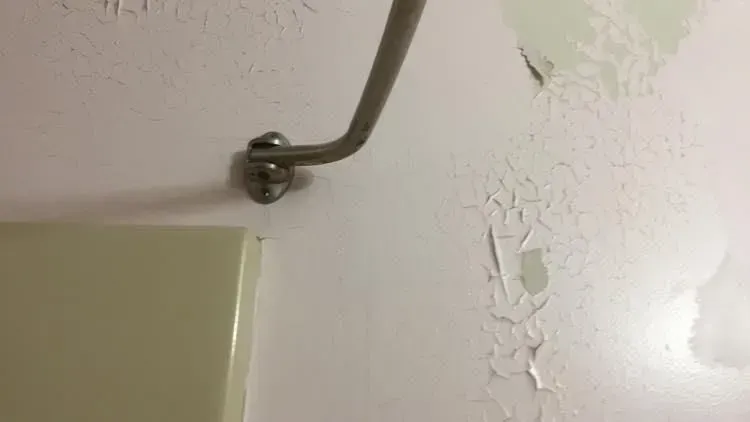 Close-up of peeling pink paint on a wall, next to a metal curved rod attached with a circular base, and the edge of a white object.