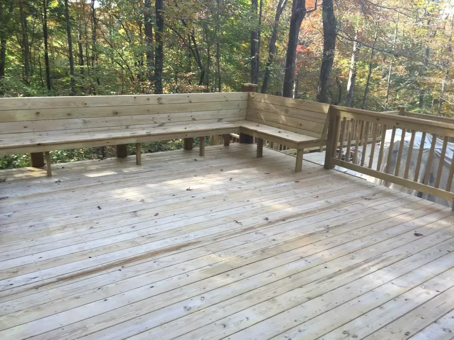 Wooden deck with a built-in L-shaped bench and railing in a wooded outdoor setting.
