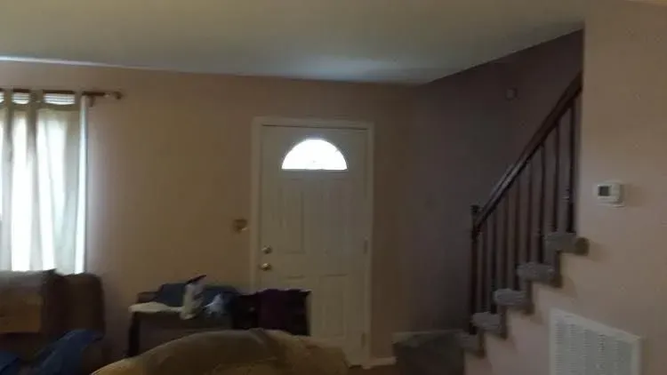 Living room with a white door, staircase, and beige walls. A couch and window with curtains are visible.