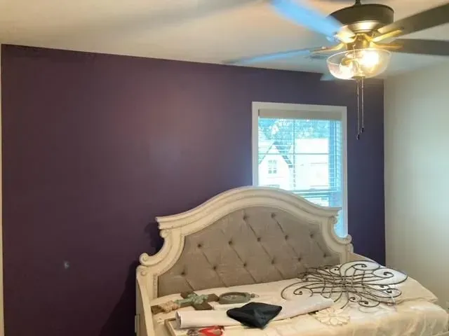 A bedroom with a purple accent wall, a white and grey upholstered headboard, and a ceiling fan.