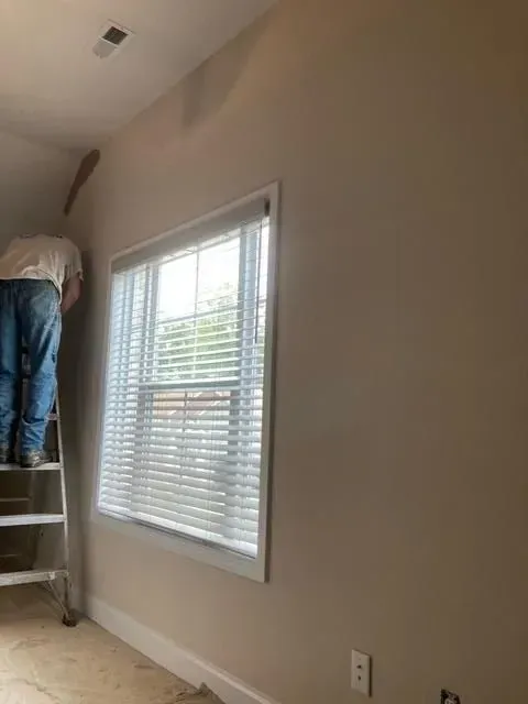 Person on a ladder painting a wall near a window with blinds. Beige walls, white window trim, and a light-colored ceiling.