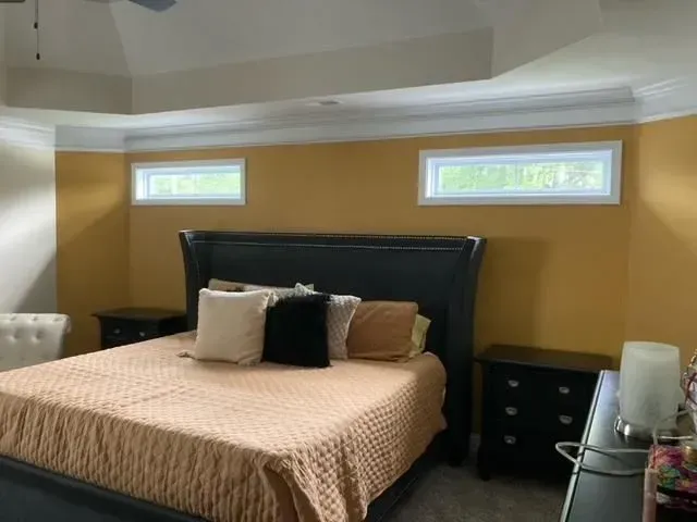 Bedroom with a bed, two nightstands, and a gold-colored accent wall with two rectangular windows. The ceiling is white with cream-colored molding.