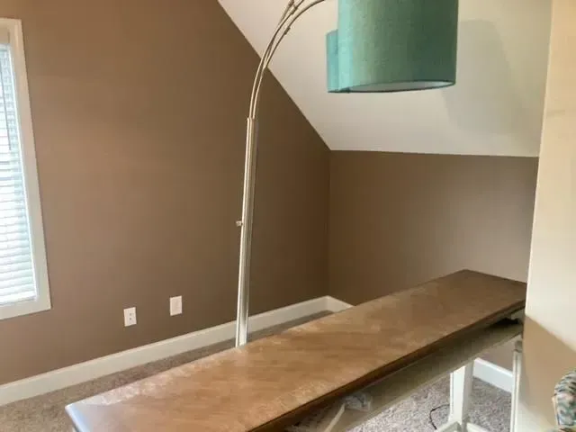 A room with brown walls, a large desk, and a silver arc lamp with a teal lampshade. A window with blinds is on the left.