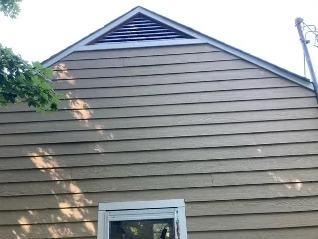 Tan house siding with a triangular vent at the top, white-framed window at the bottom.