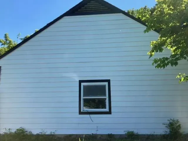 Side view of a white building with a black-framed window and roof. The sky is blue with tree branches visible on the right.