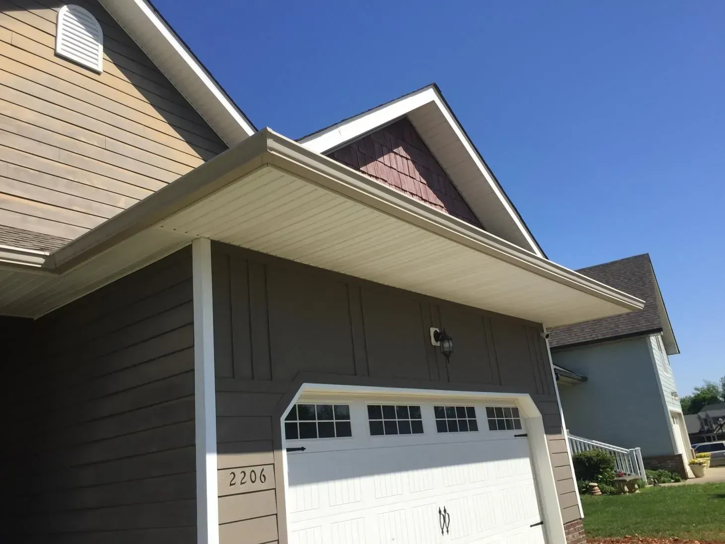 Garage exterior with brown siding, white garage door, and a beige roof overhang. Blue sky in the background.