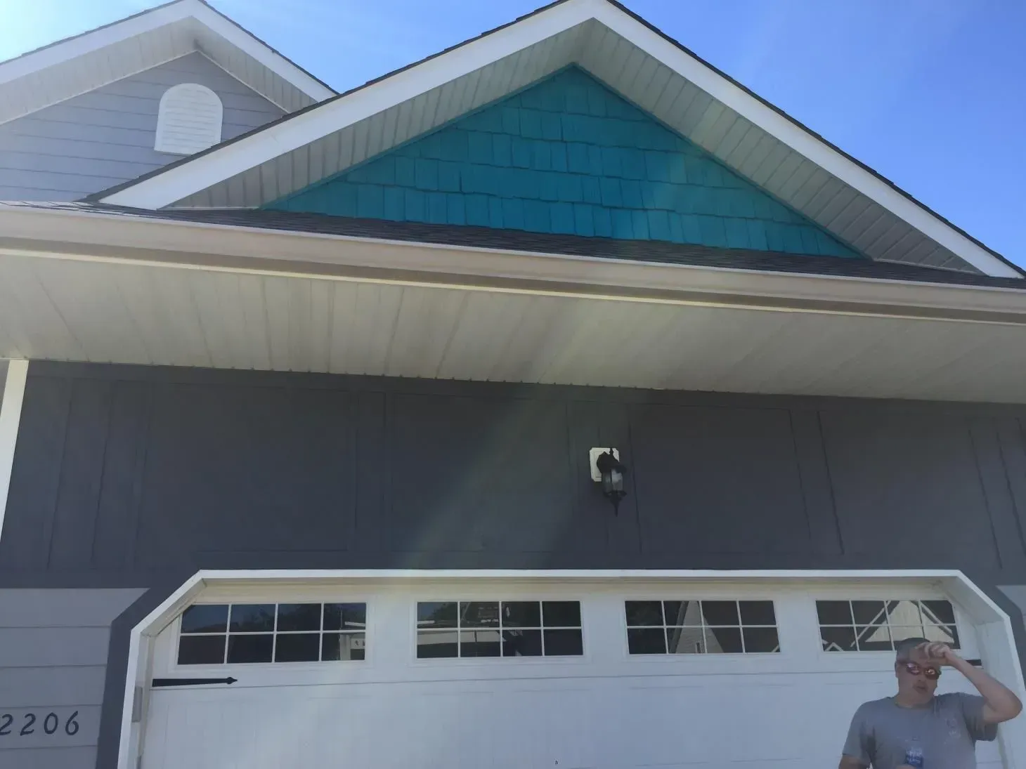 Gray house with white garage door; teal-colored gable. A person stands in front, looking upward.