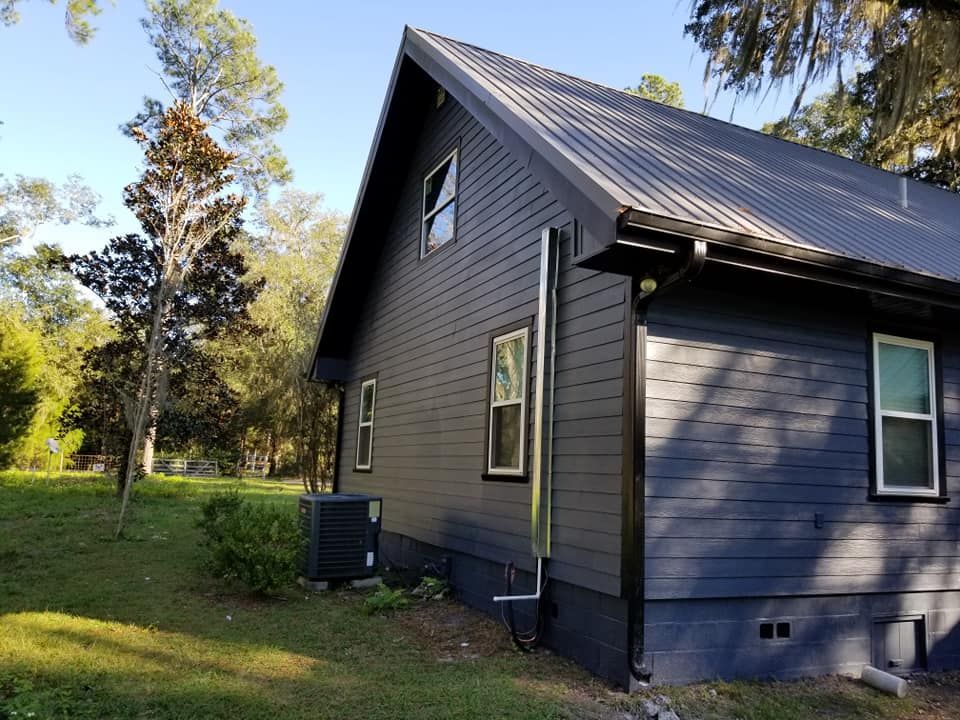 A dark blue house with a metal roof sits in a grassy area. Several windows and a metal chimney are visible.