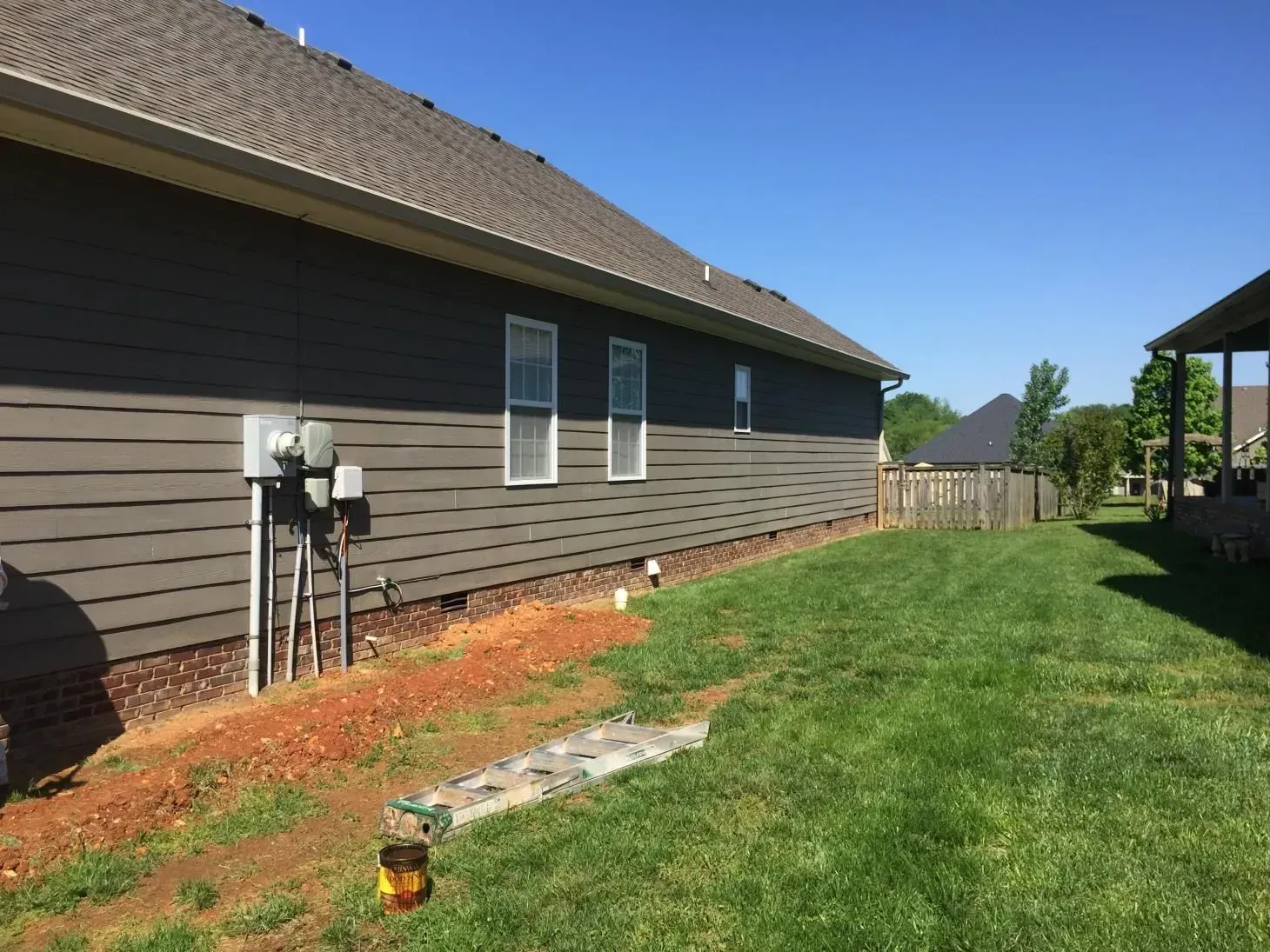 Side of a house with gray siding, windows, and a grassy yard. A bare dirt patch and electrical boxes are also visible.