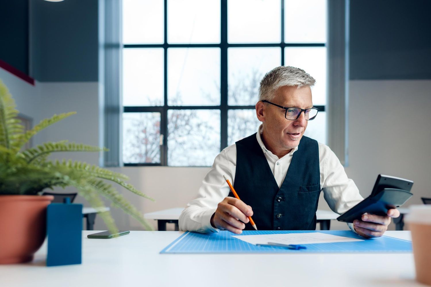 Man in glasses uses a calculator, working at a desk, near a window and a plant.