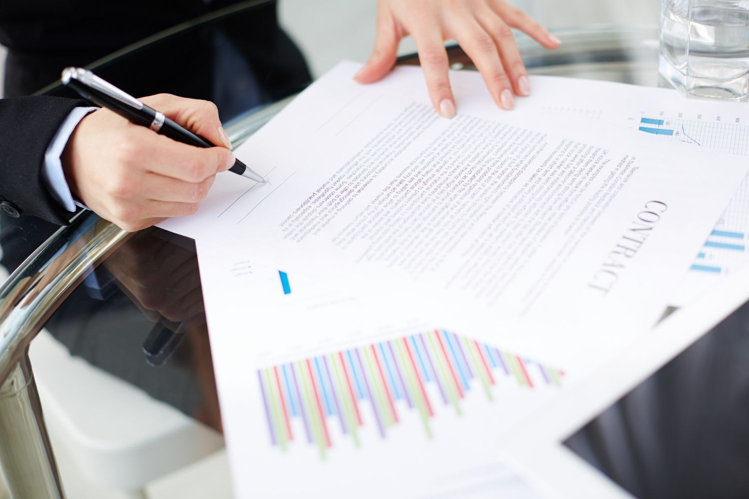 Person signing a contract at a table with charts and a glass of water.