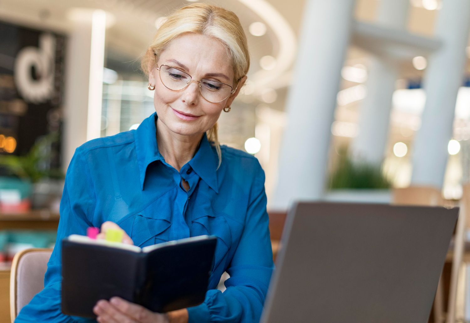 Woman with glasses, blue shirt, looking at a notebook, sitting near a laptop, indoors.