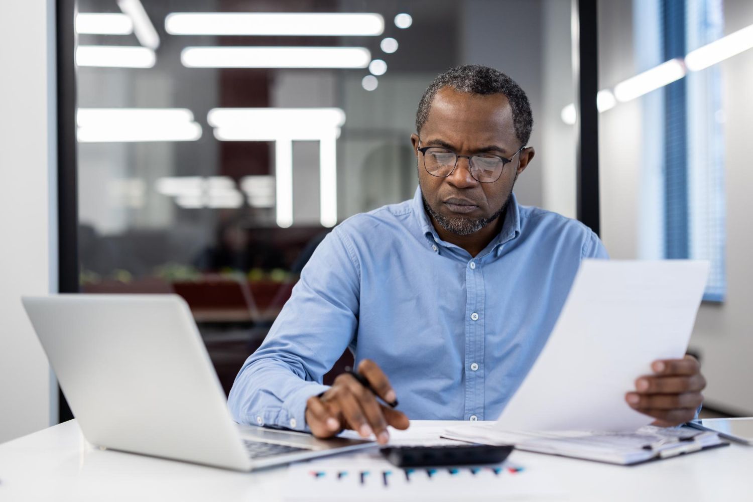 Man in blue shirt, glasses, working on laptop, calculating with documents in office setting.