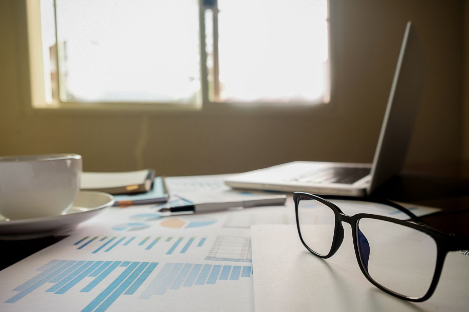 Laptop, documents with charts, glasses, and coffee cup on a desk near a window.