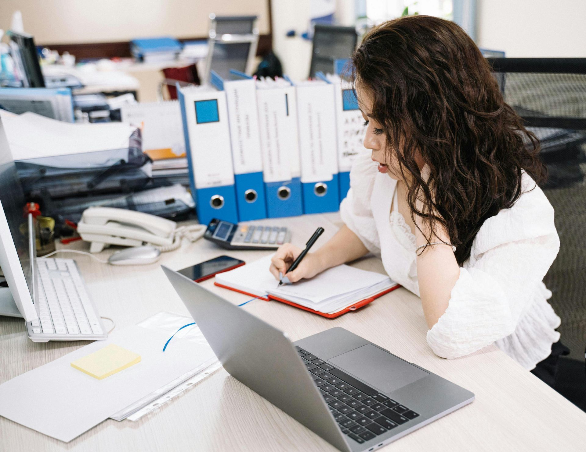 Woman writing in a notebook at a desk with a laptop, calculator, and papers in an office setting.