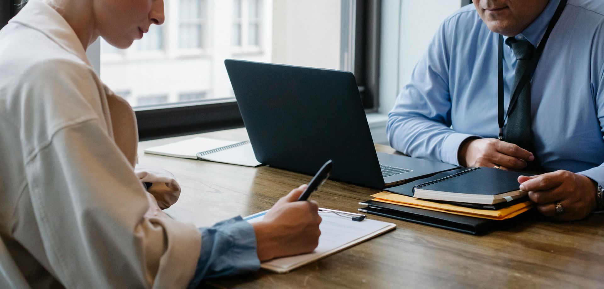 A person writes on a clipboard at a desk while another sits opposite, a laptop and notepad in view.