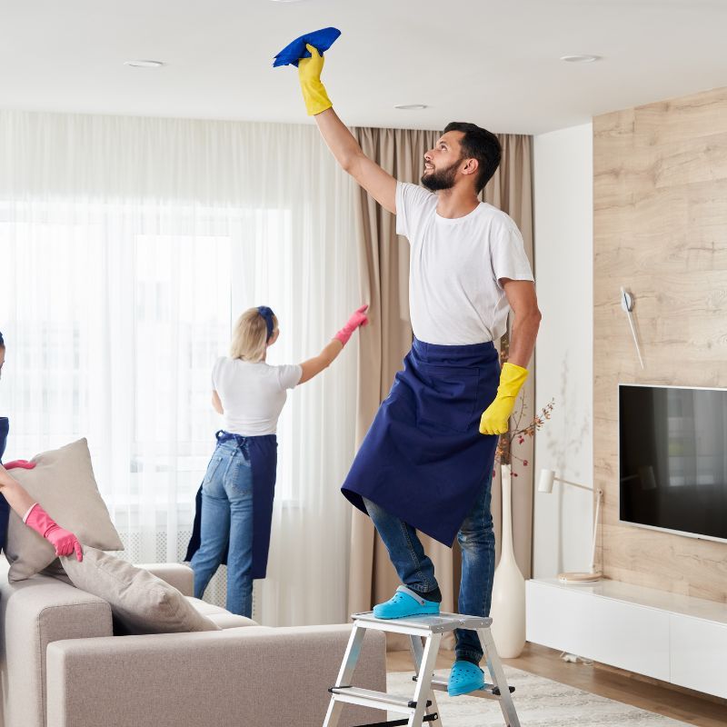 Three people cleaning a living room: one on a ladder wiping the ceiling, two others cleaning furniture.