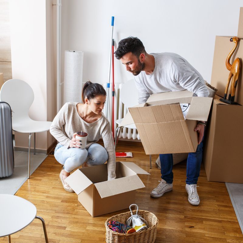 Couple unpacking boxes in a room with hardwood floors, moving furniture, and holding items.