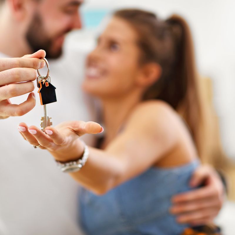 Man handing keys to woman, both smiling and embracing. The keys have a house-shaped keychain.