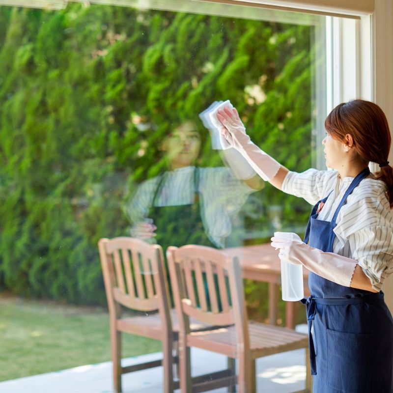 Woman in apron and gloves cleaning window. A wooden table and chairs visible outside.