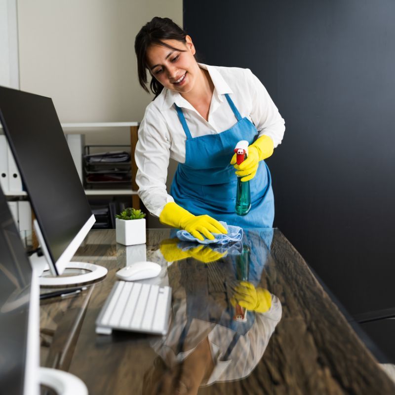 Woman in blue apron, gloves, sprays and wipes desk with a smile in an office.