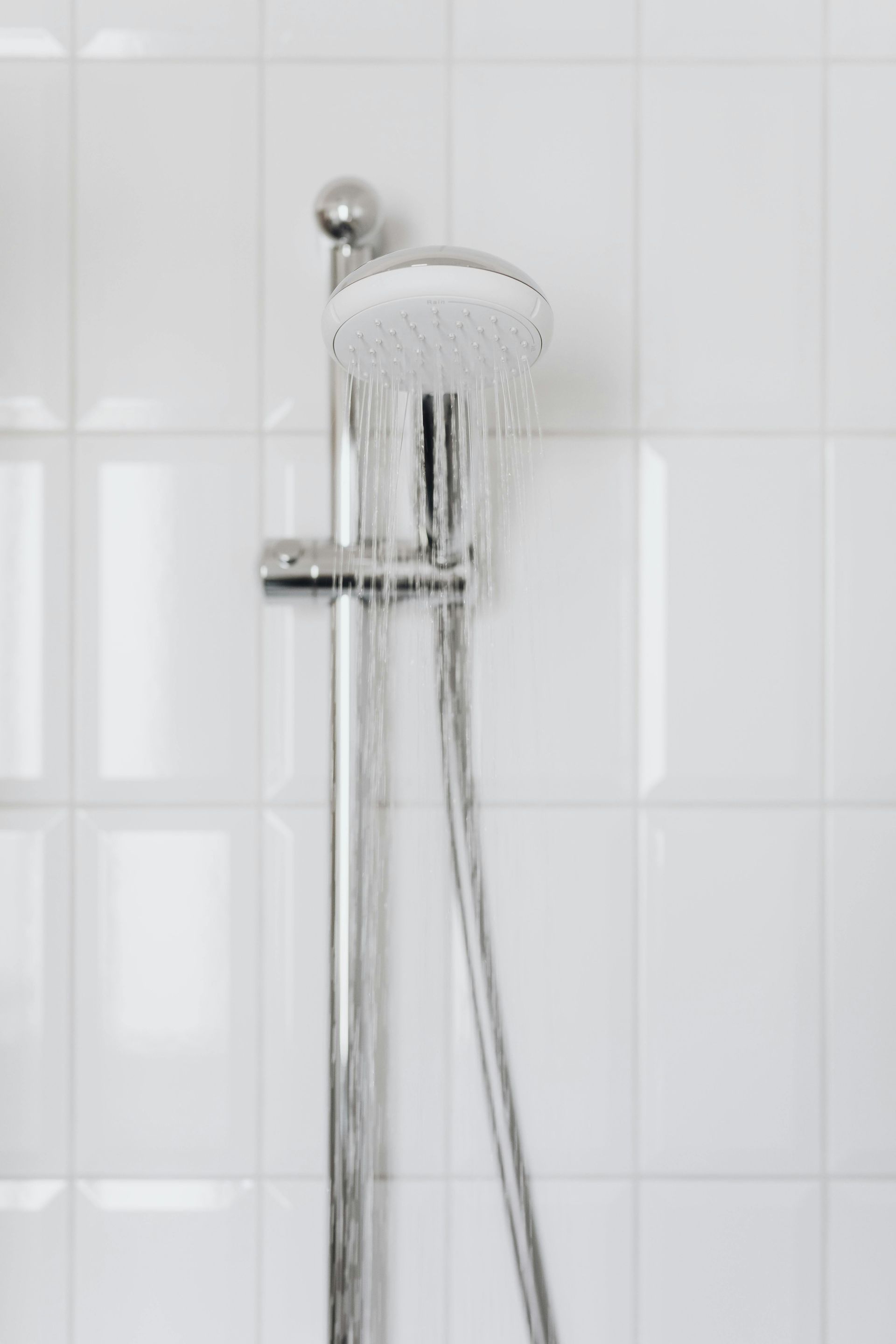 Showerhead with water spraying, against white tiled wall. Chrome fixtures, minimalist bathroom design.