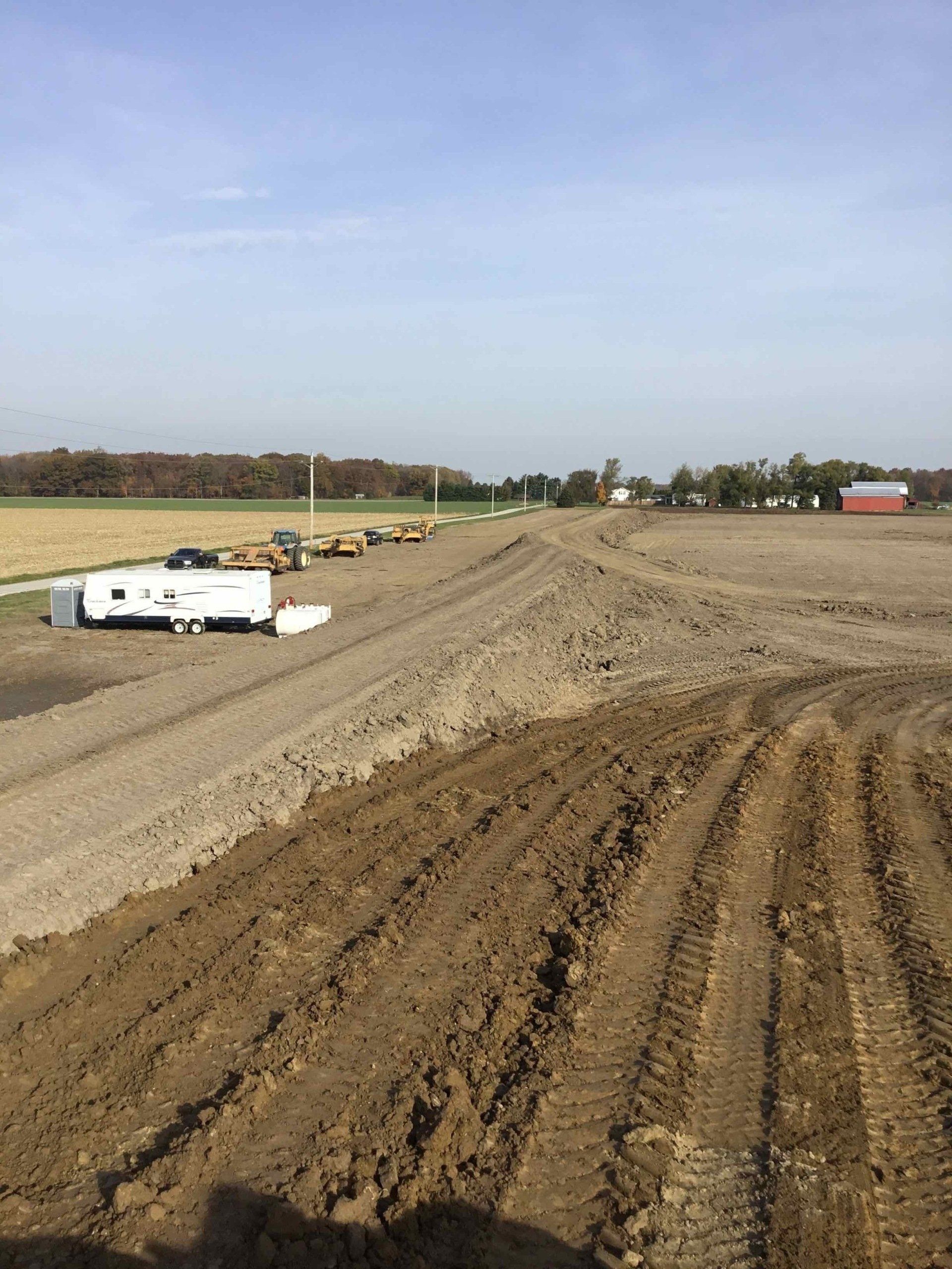 Dirt path being built, with a line of construction vehicles in the background. Field, trees, and sky.