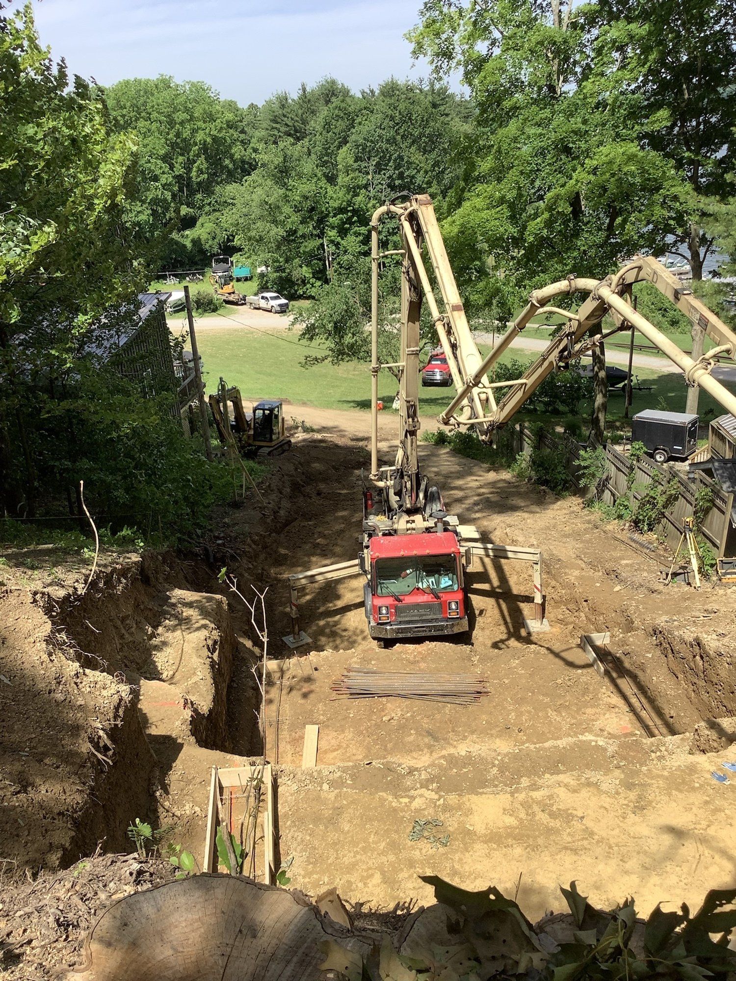 Construction site: red cement truck with pump pouring concrete into a trench. Yellow pump arm extends over the site. Trees in background.